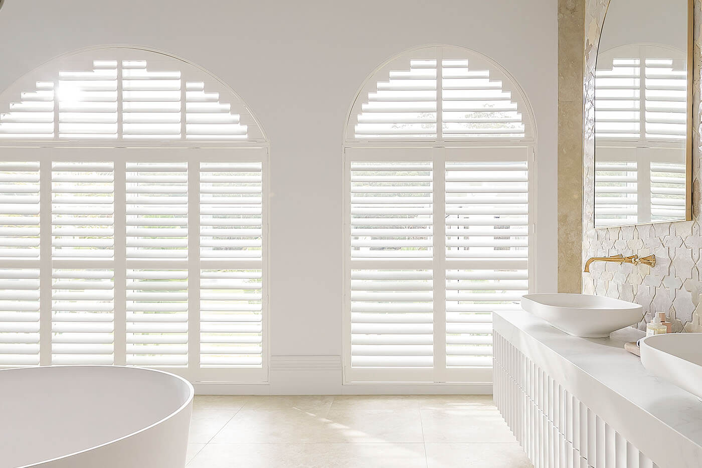 Classic transitional style white bathroom with large radius windows covered by Polysatin Shutters, offering light and privacy control in the bathroom. Suitable for wet areas. For sale in our Brisbane showroom.