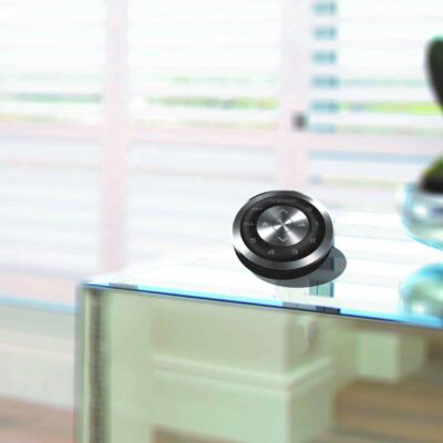 A sleek black remote Norman curtains and blinds control device sits on a modern glass table with a blurred background of white plantation shutters and wooden flooring. A plant in a glass bowl is partially visible on the right side of the image.