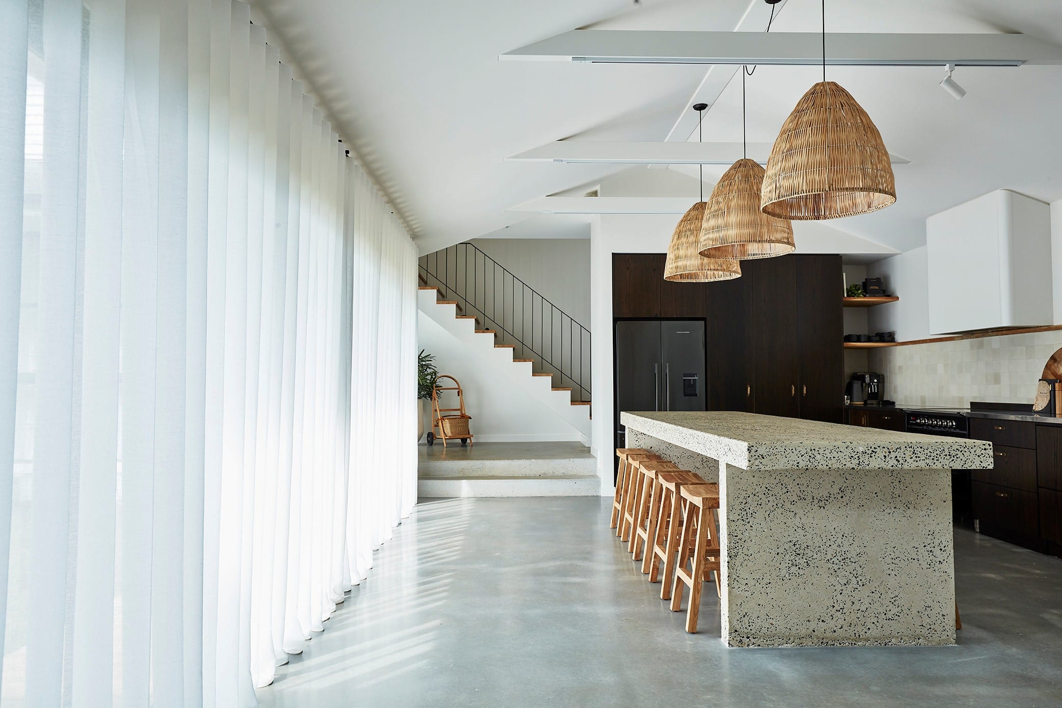 Modern minimalist kitchen with a large terrazzo island, wooden bar stools, and woven pendant lights. The space features polished concrete floors, dark cabinetry, and a staircase with a sleek black railing. Floor-to-ceiling sheer white curtains by Complete Blinds line the left side, allowing natural light to fill the room.